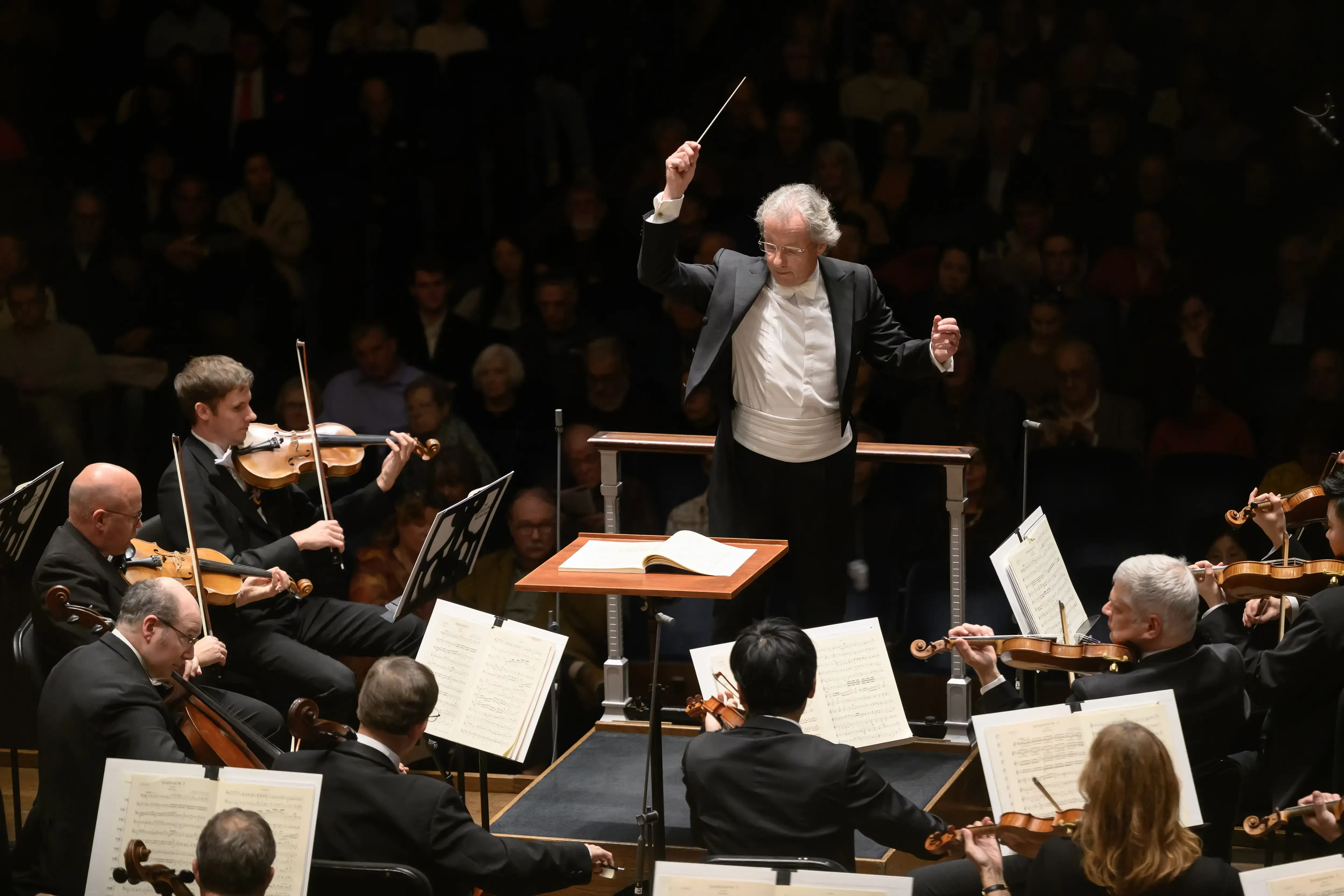 Franz Welser-Möst leads an orchestra with string musicians performing on stage, surrounded by an audience in a dimly lit Mandel Hall.