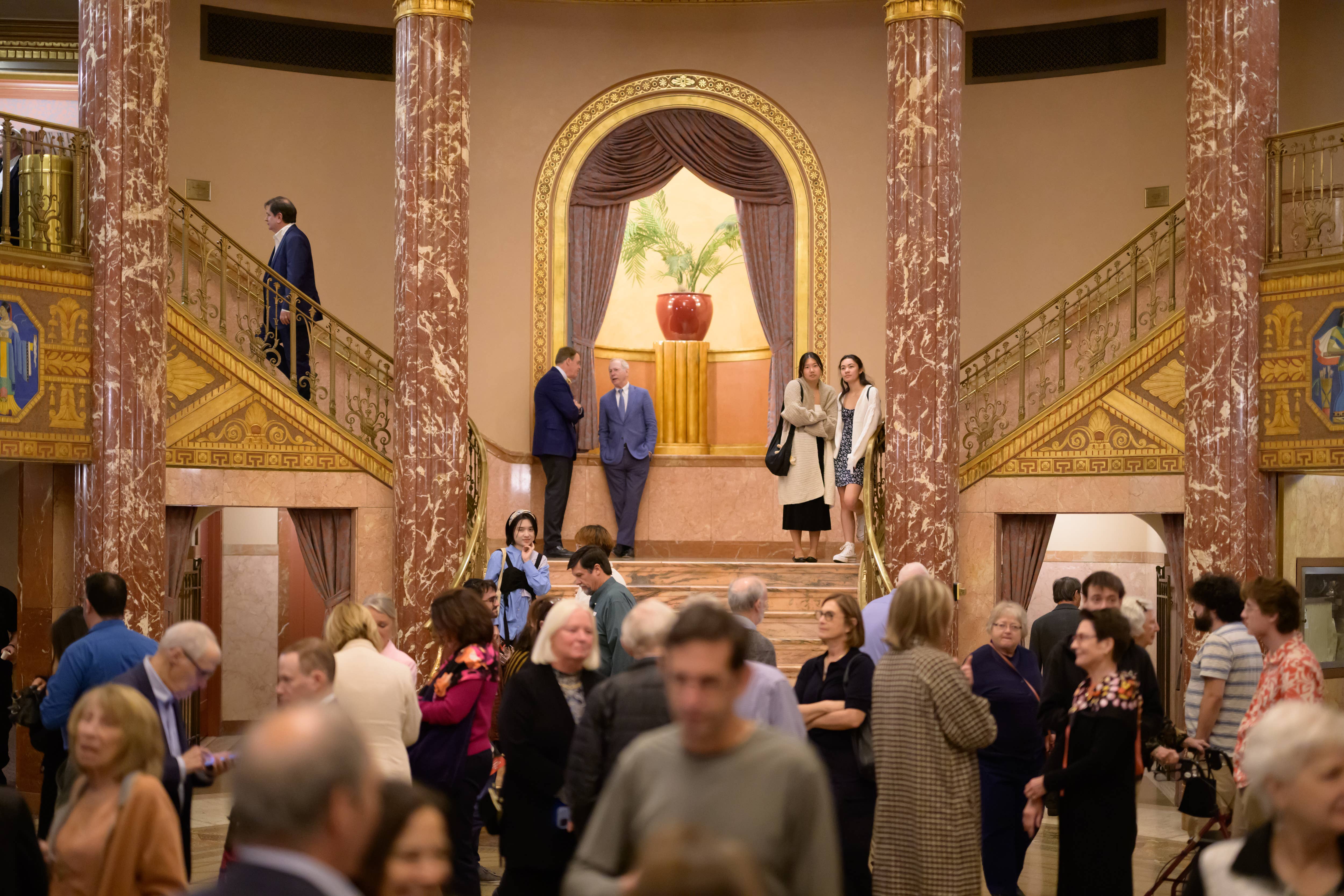 Elegant Grand Foyer with marble columns, ornate staircases, and people mingling. A red vase is displayed on a pedestal.