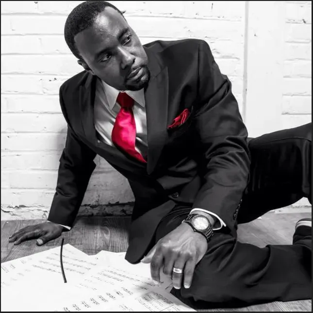 Black-and-white portrait of a man in a dark suit with a red tie, leaning on a desk by a brick wall.