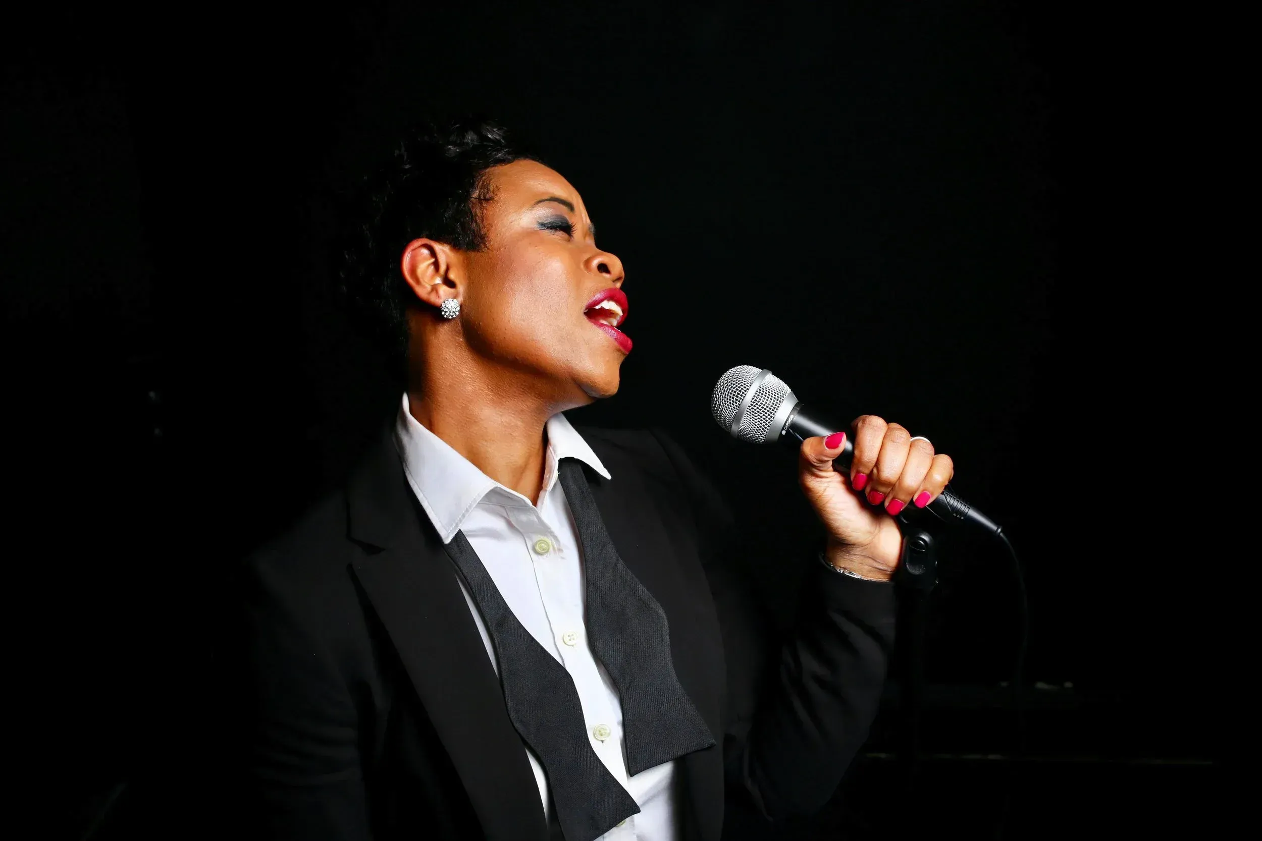 The Phoenix in a blazer singing into a handheld microphone against a dark background.