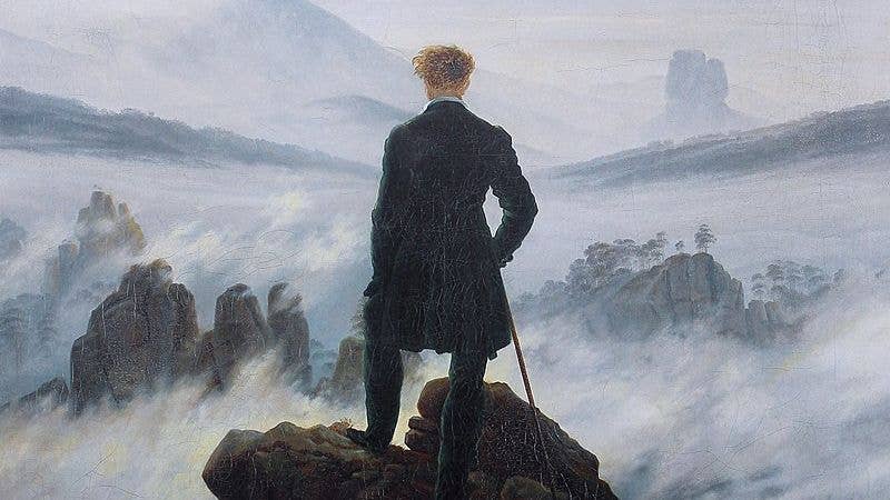 Man in black with walking cane stands upon a craggy height just above the fog. Looking away from us, he gazes upon the seemingly endless mist-shrouded landscape before him