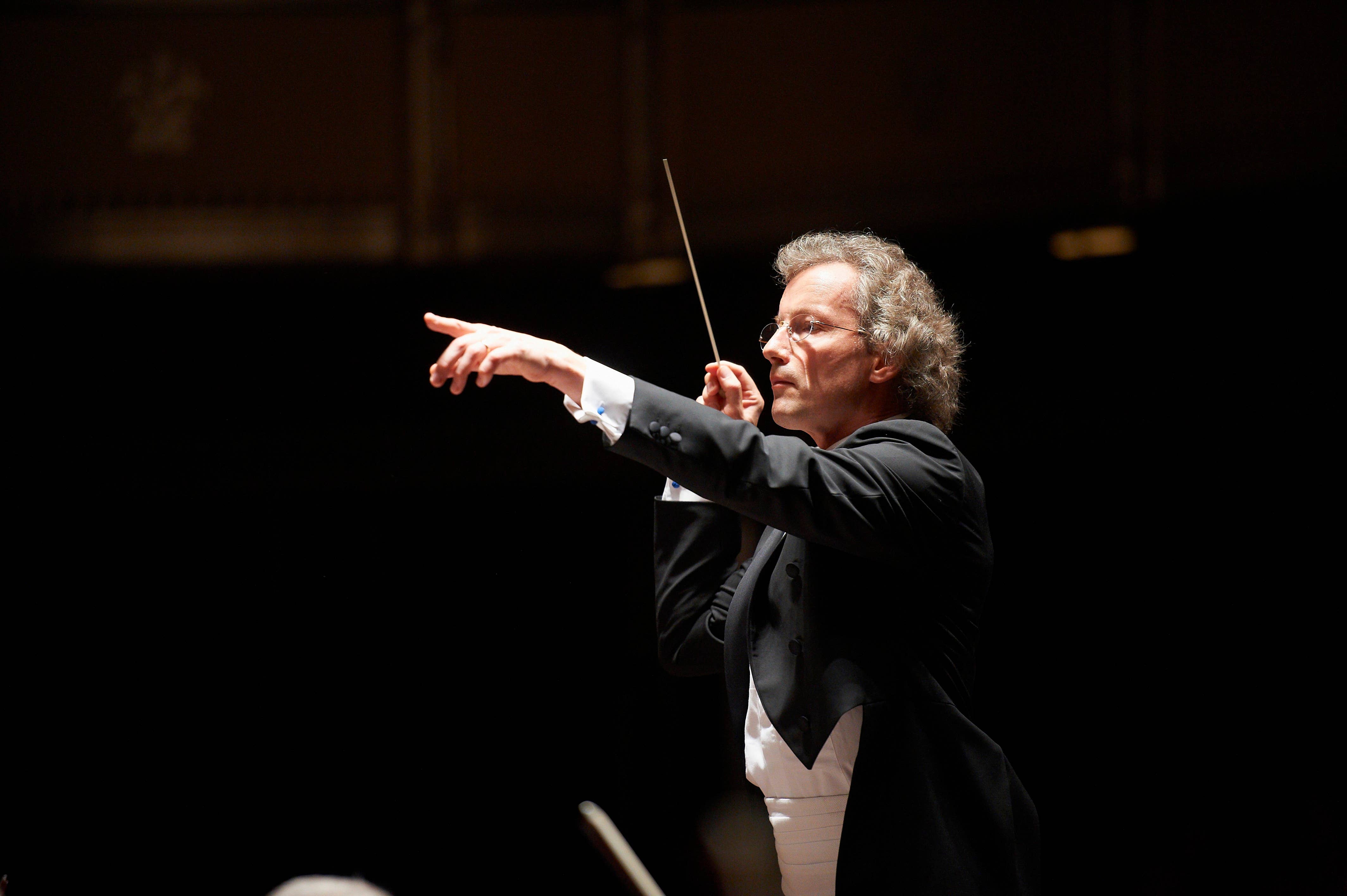 Against a dark backdrop, the music director is illuminated in a dramatic conducting pose.