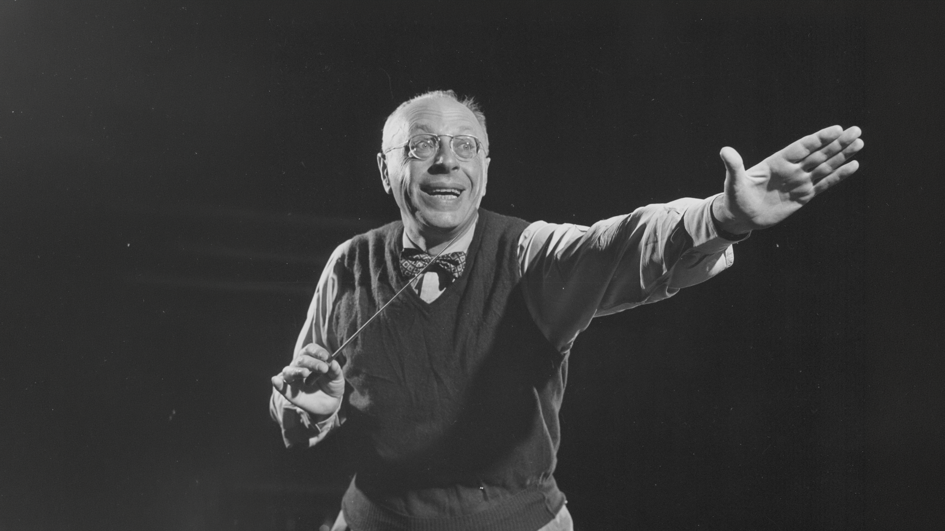 George Szell dressed in a sweater vest. Smiling, he excitedly points forward with his left hand. His right hand holds his baton.