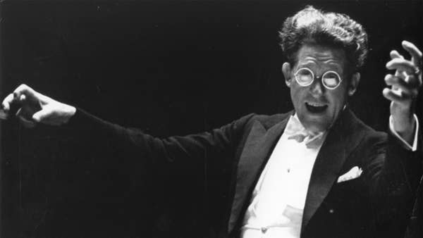 Artur Rodzinski conducting the Orchestra. He is facing the viewer, has a huge, excited smile on his face, and is wearing large glasses.