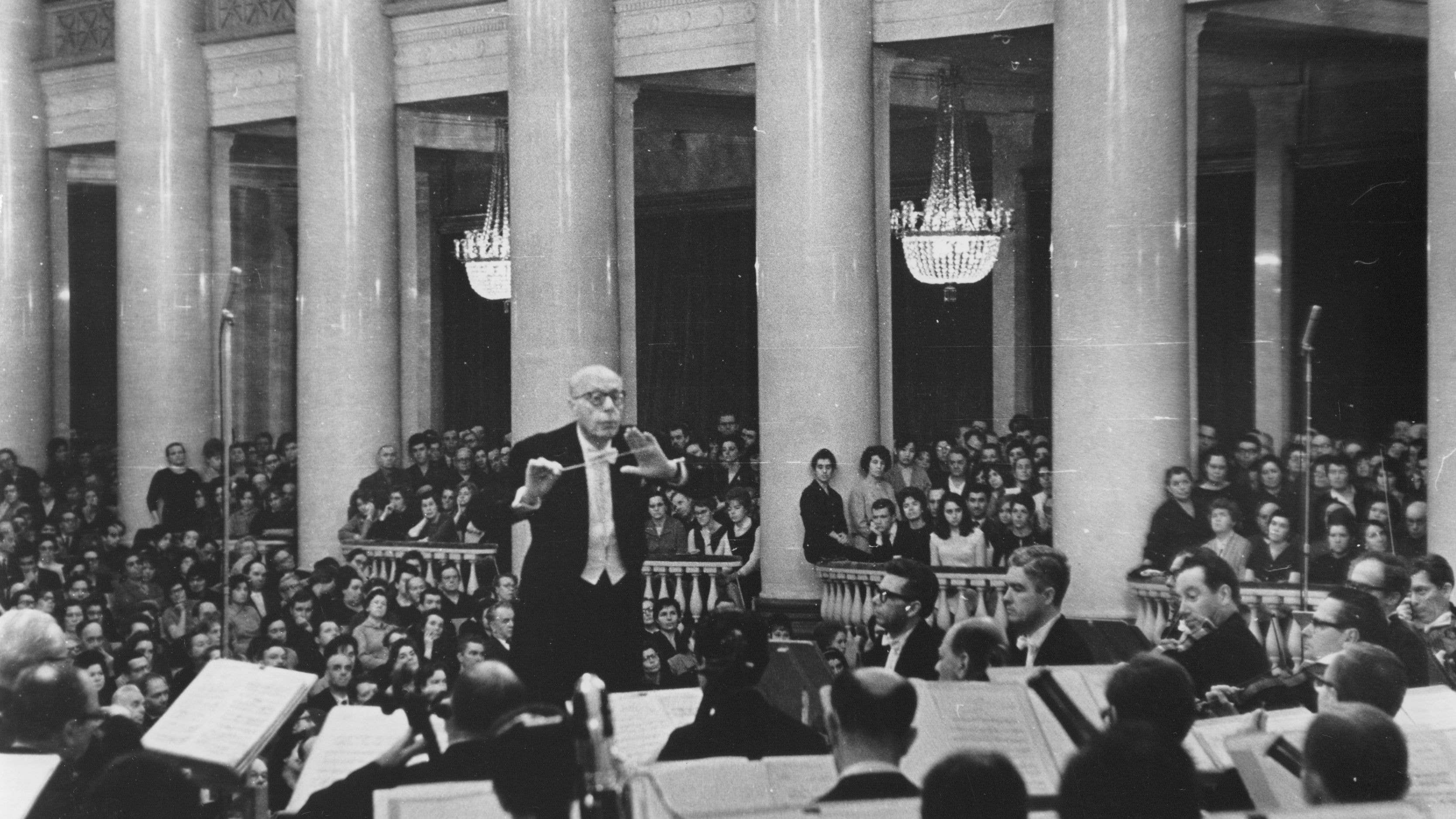 Szell is conducting the Orchestra. A large, attentive audience is seated behind them. Prominent chandeliers hang above the audience.