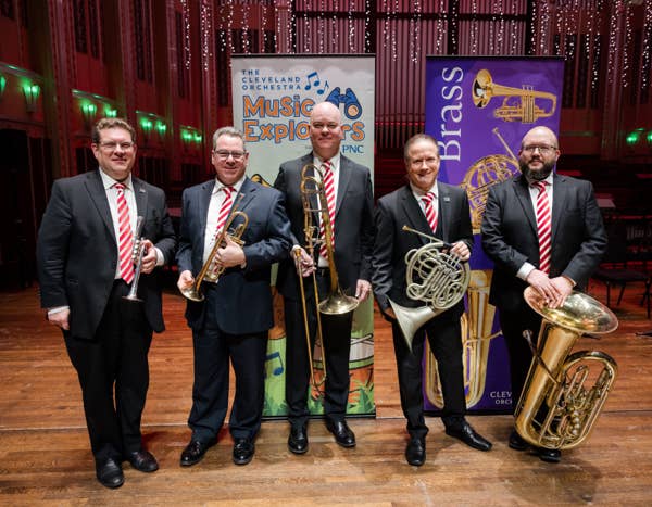 Five men in suits holding brass instruments in a theater lobby with concert banners and lit stage backdrop.