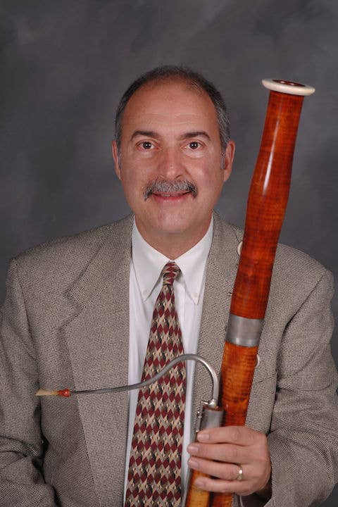 Mark DeMio in suit holding a tall wooden instrument with a metal mouthpiece against a studio backdrop