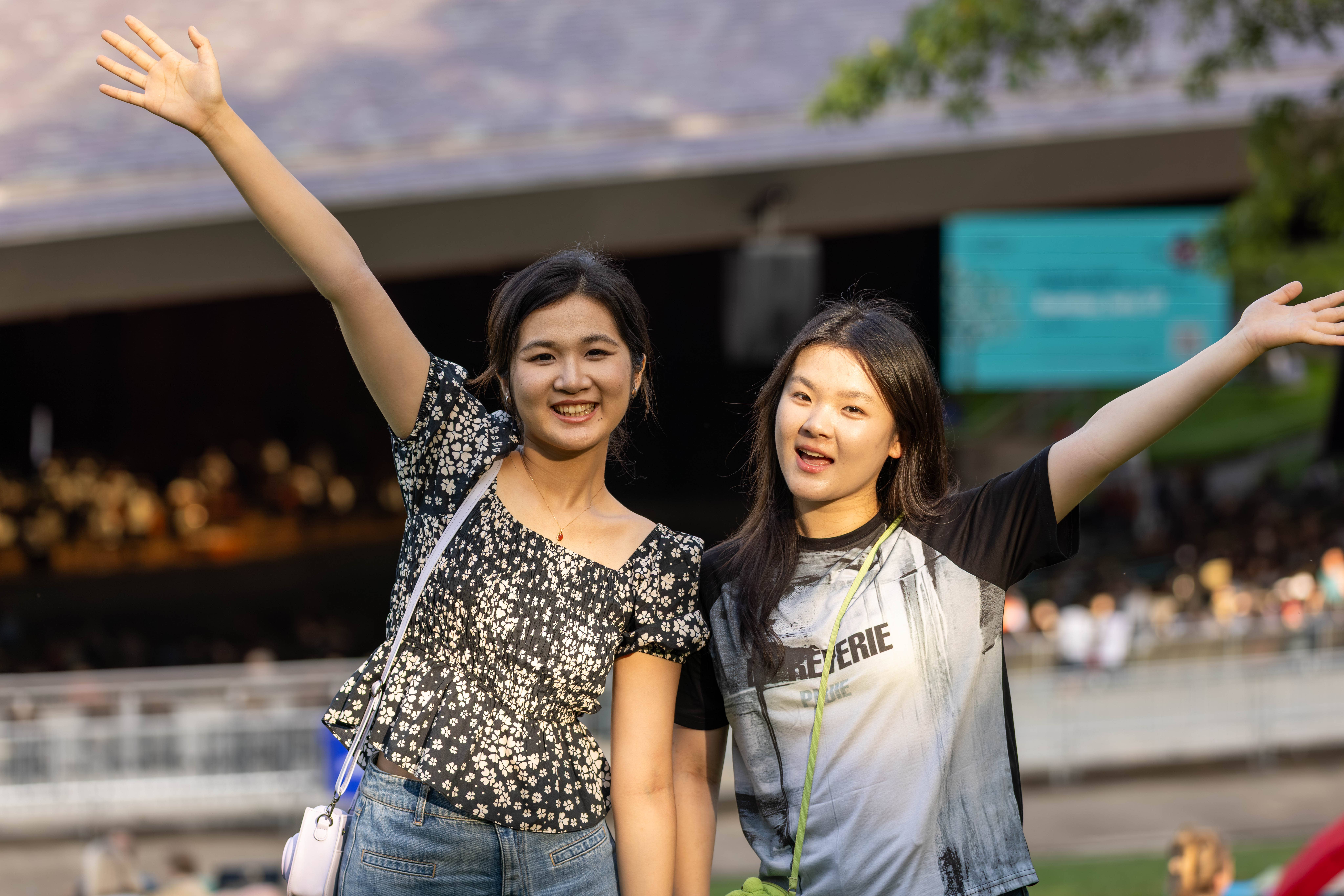Two women smile with arms raised outdoors near a pool and building in sunny weather.