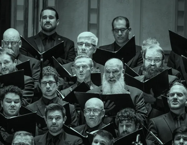 A men's choir performs, holding sheet music and wearing dark suits, with expressions of focus and intensity.