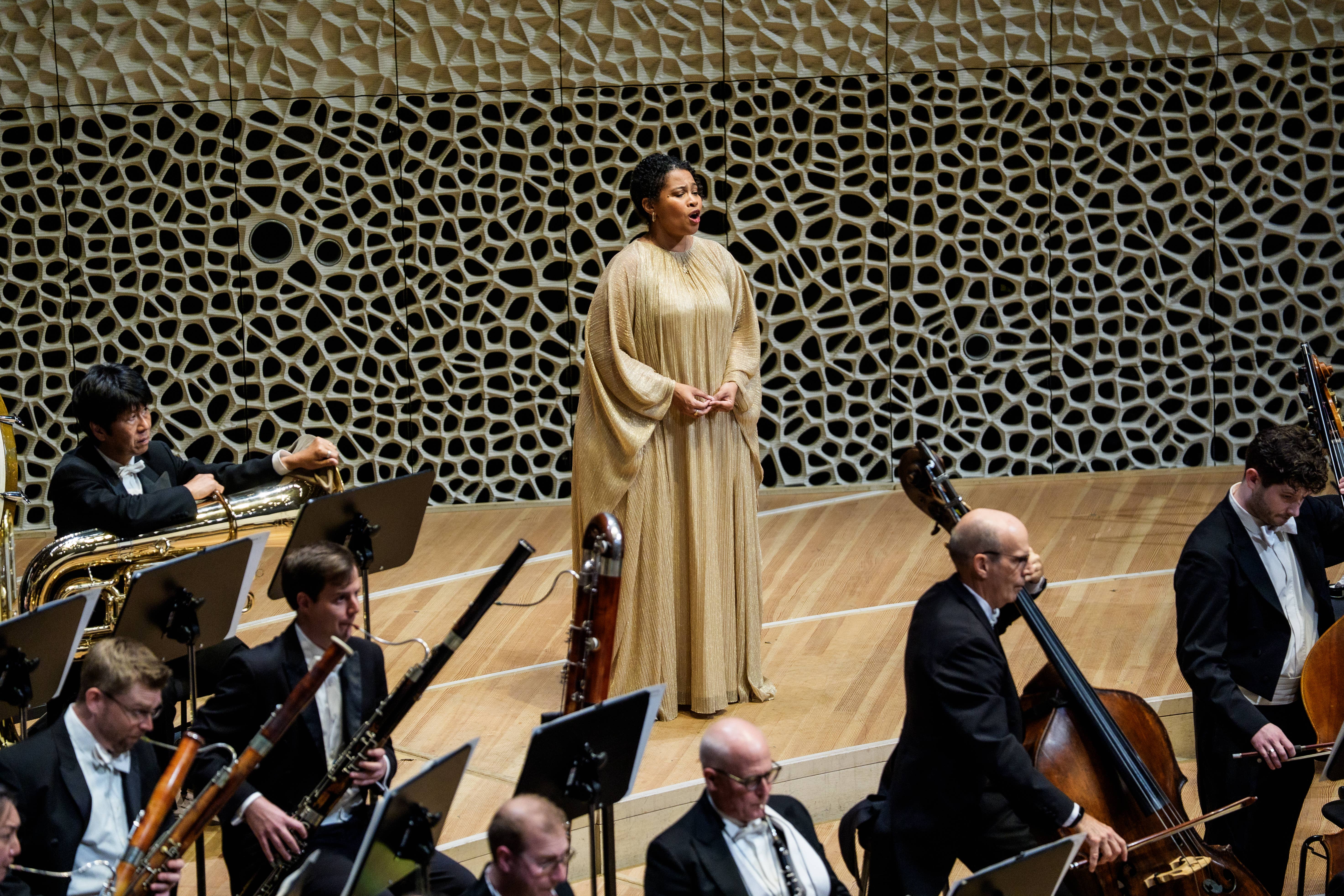 A woman in a gold dress sings behind an orchestra