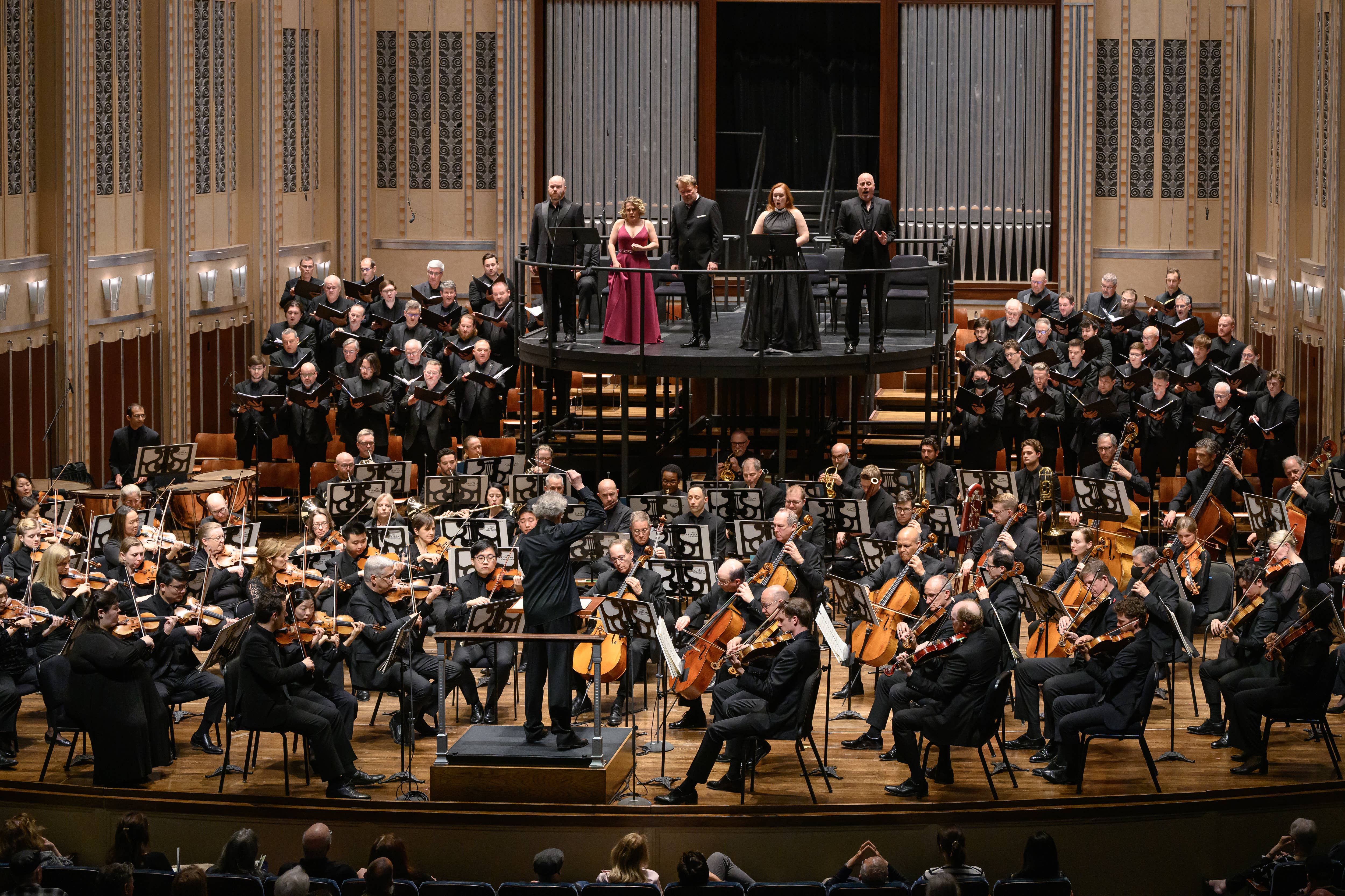 Orchestra seated onstage with conductor on podium and solo performers singing under ornate hall backdrop