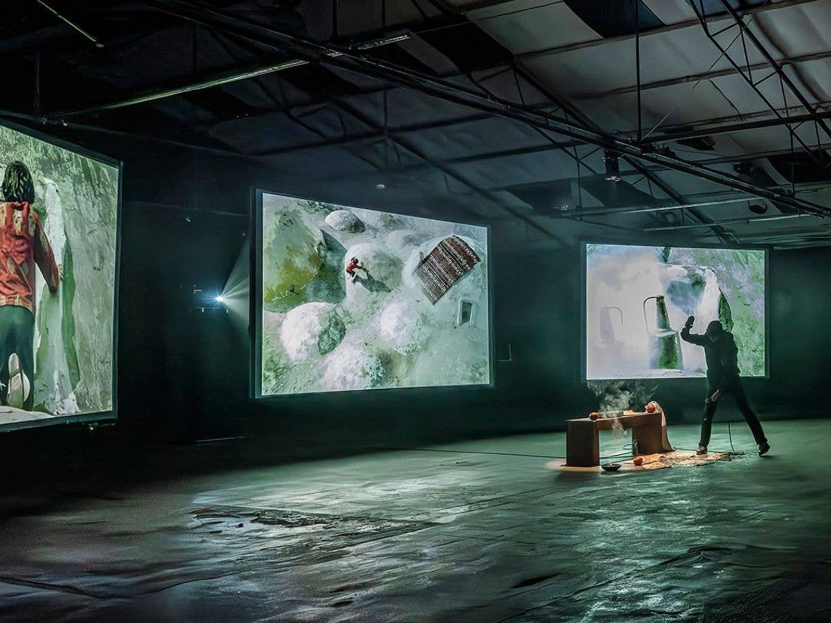 Audience silhouettes watch a performance projected on three large screens in a dark gallery space.