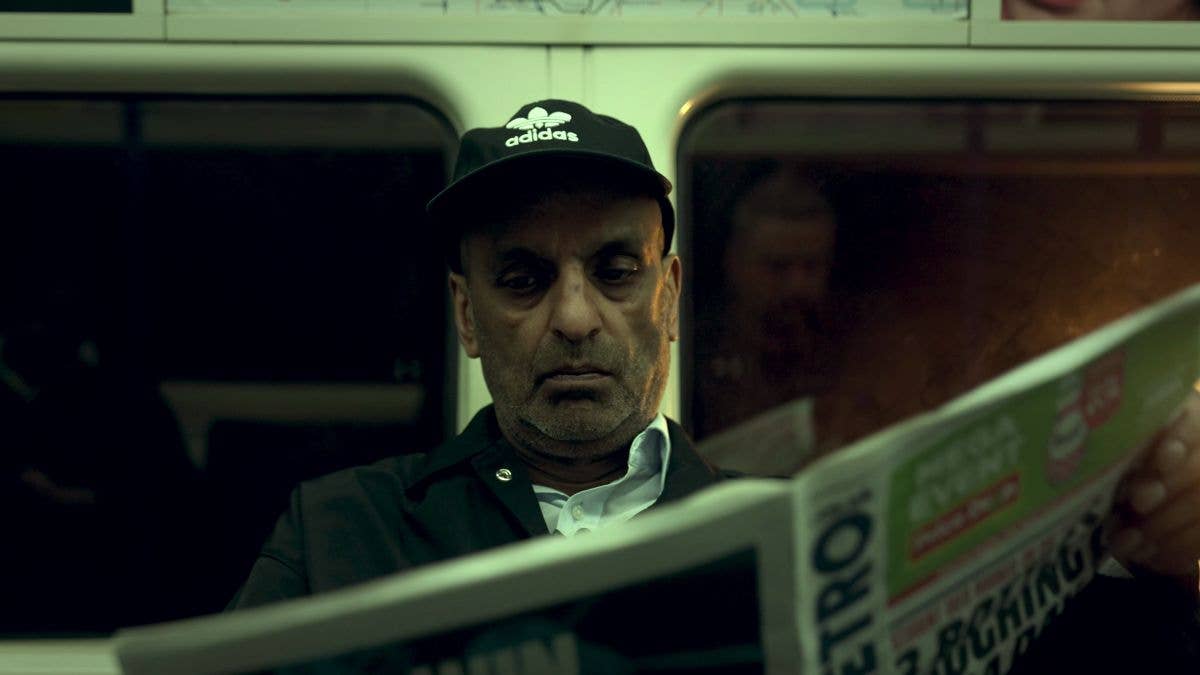 Satpal wears a black Adidas cap holding a newspaper while sitting on public transport, seen through a window.