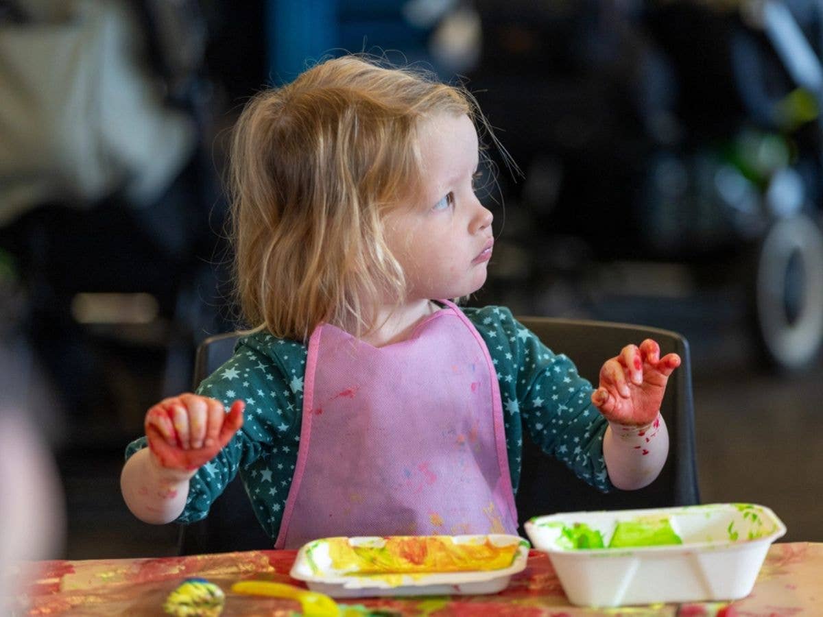 A young child sits at a table with a pink bib, holding up their hands which are covered in paint.