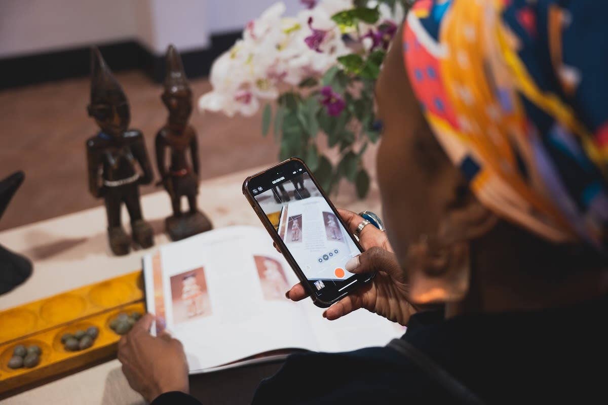An over the shoulder shot of someone taking a photograph of the open pages of a book on their phone. The book contains images of sculpture.