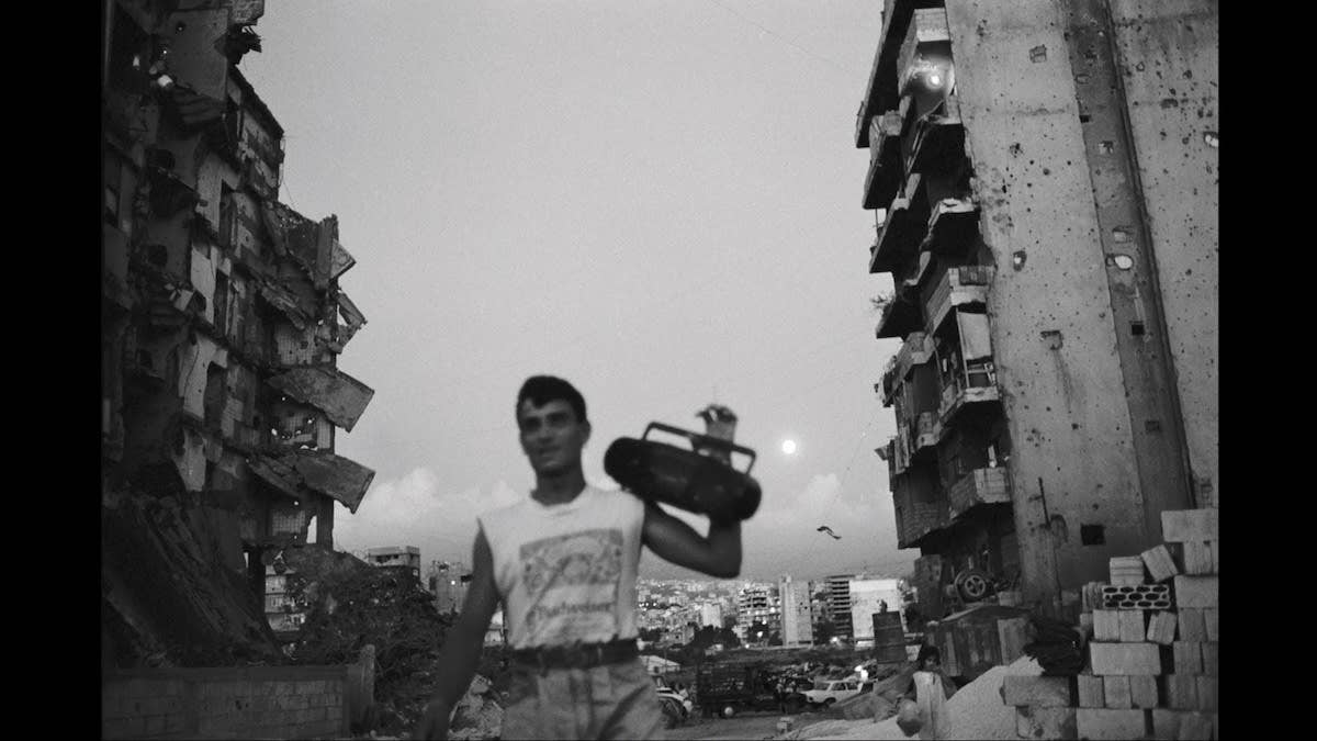 Still from Do You Love Me. The sun is setting as a person walks holding a CD player on their shoulder with ruined buildings in the background.