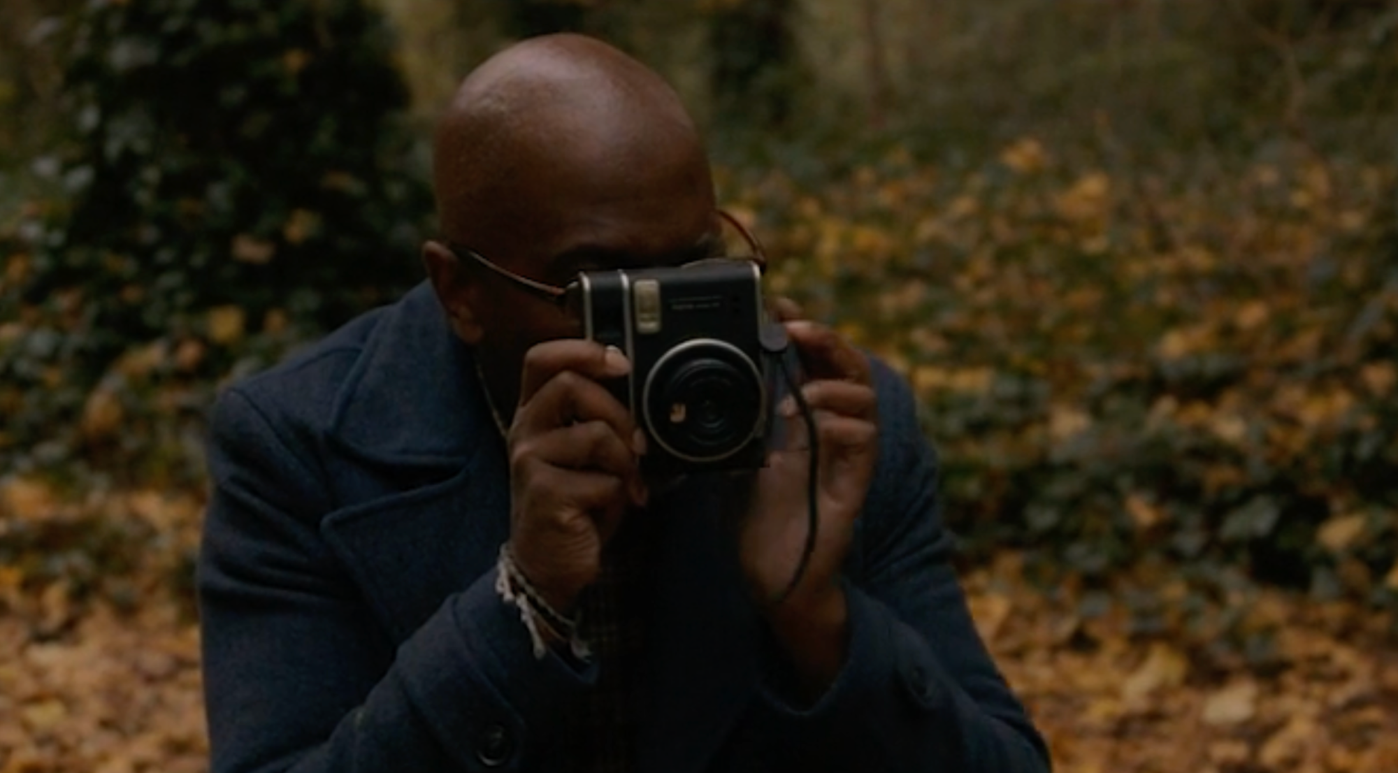 A photographer mid-shot, holding the camera up, obscuring their face. They are surrounded by nature - trees and leaves.