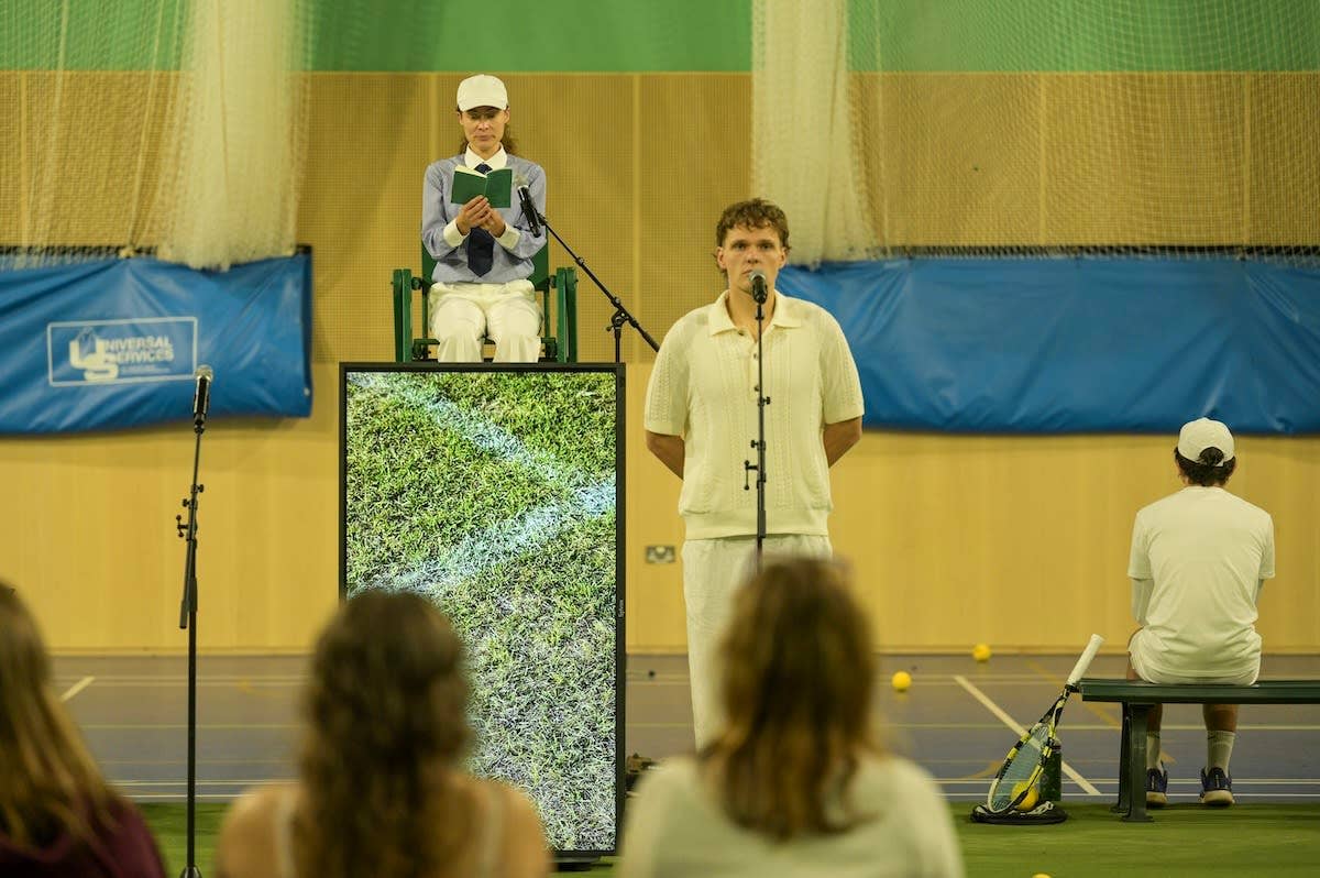 A person in tennis whites stands before a microphone, with an official in a cap behind them looking at an open book.