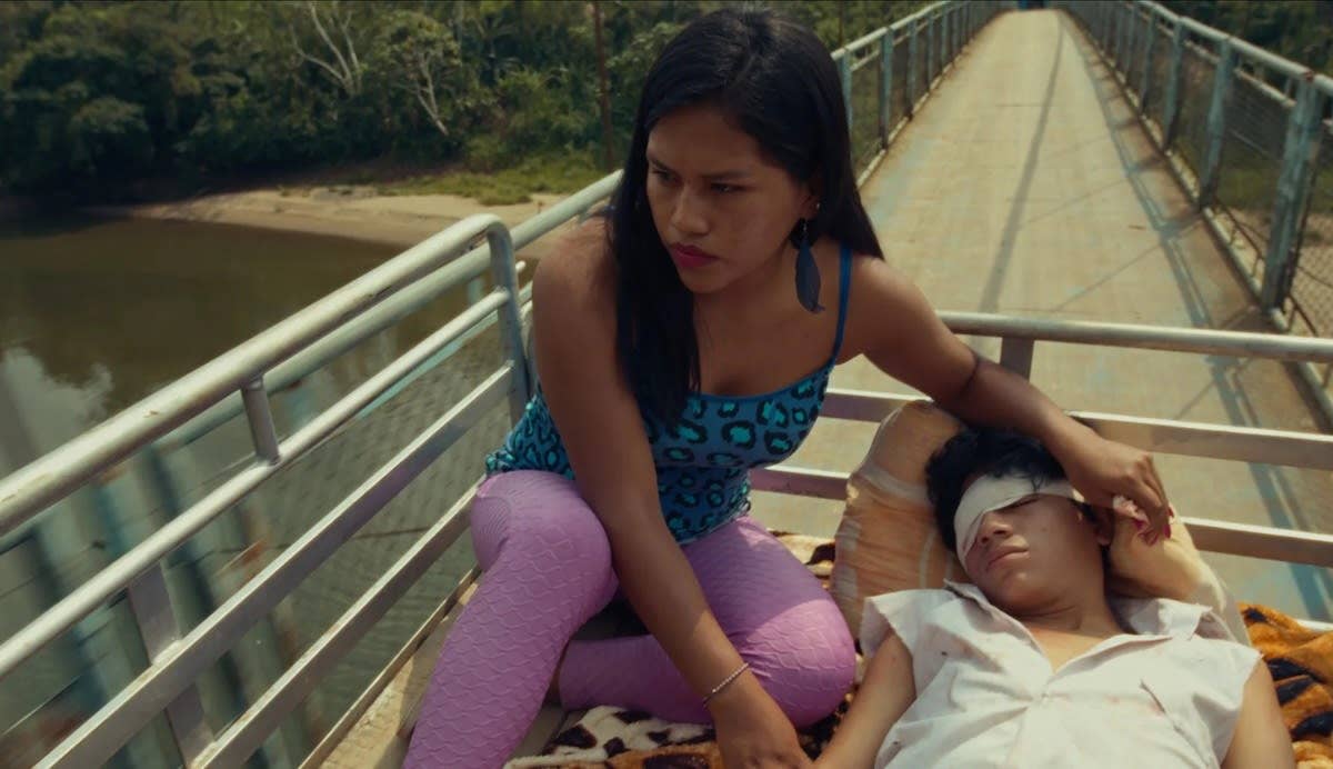 Characters Meshia and Iván travelling in the back of a truck, Meshia's hand rests protectively over Iván whose eye is bandaged.