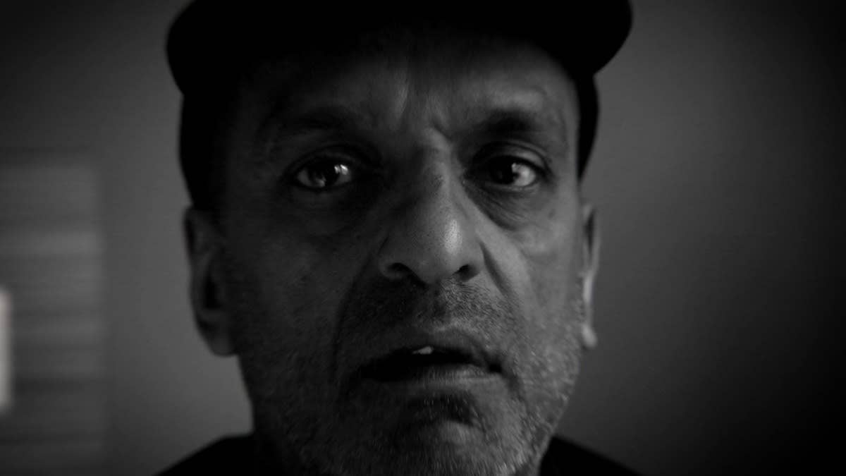 A black and white close up of Satpal Ram's face, looking directly at the viewer.