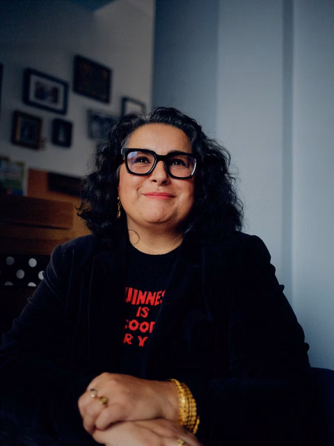 Shaheen Baig wearing glasses, a dark jacket and gold jewellery, smiling in a warmly lit room, seated at a desk.