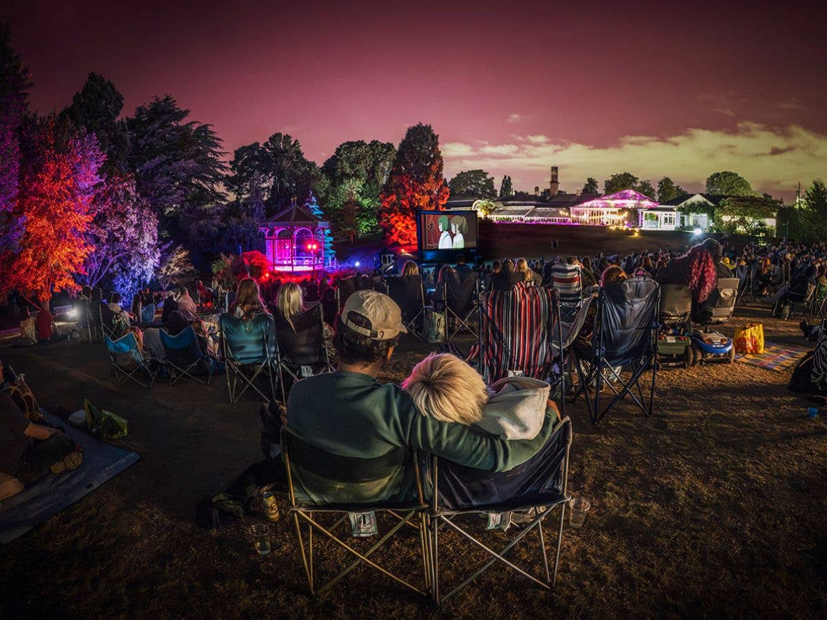 An audience sits on camping chairs and blankets, with their back towards the camera, watching Spirited Away on a large screen surrounded by lit trees.