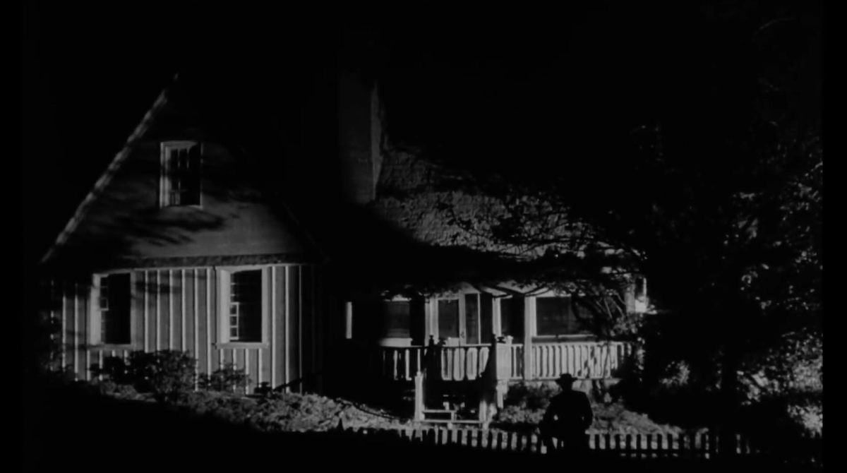 Black-and-white house at night with a shadowy figure stood in the foreground.
