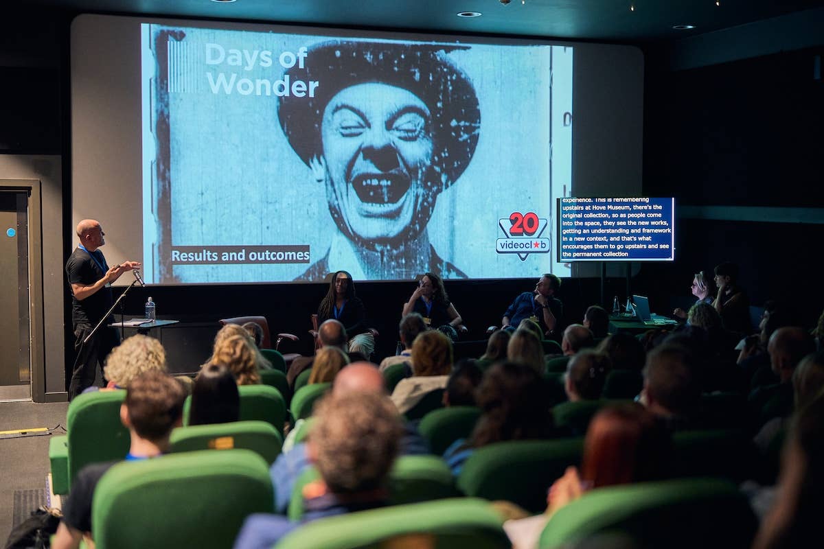 Speaker presenting on results and outcomes from videoclub's “Days of Wonder” project to an audience seated in rows.
