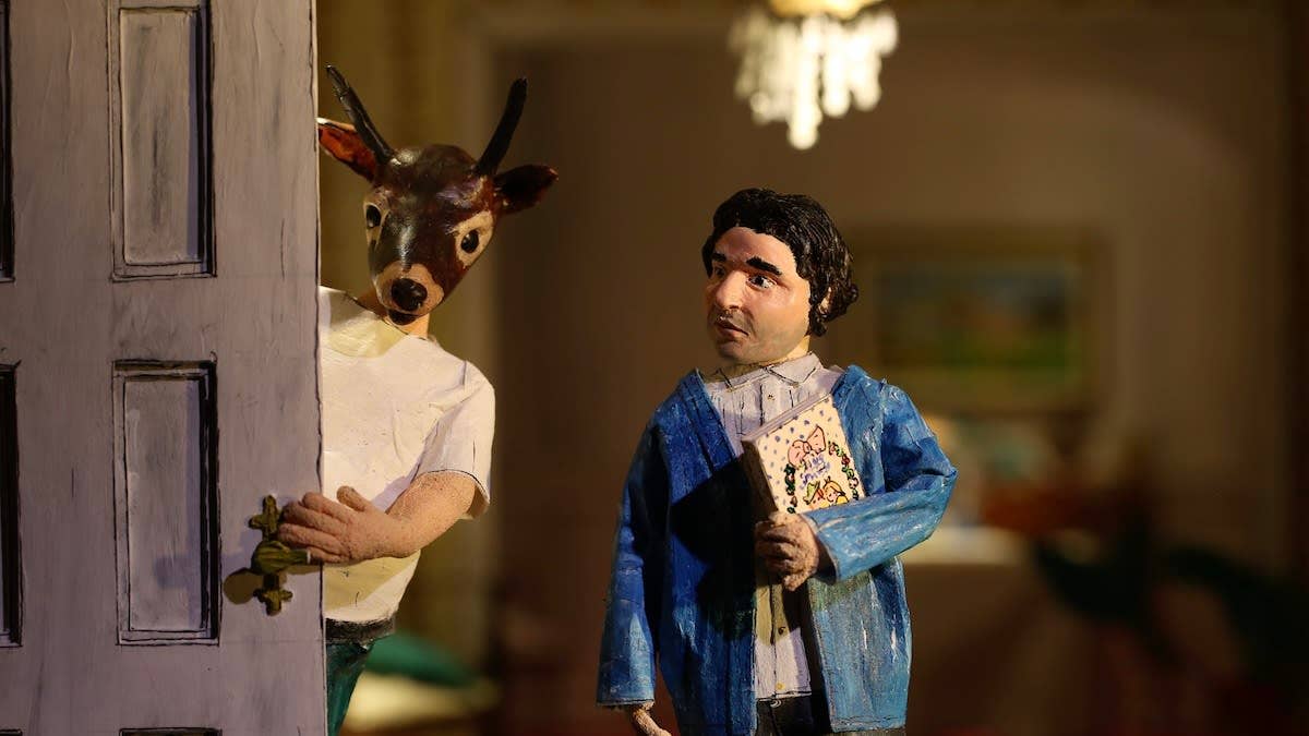 A stop motion animation scene with two figures - one with a blue jacket clutching a book, one with a deer's head peering around a door.