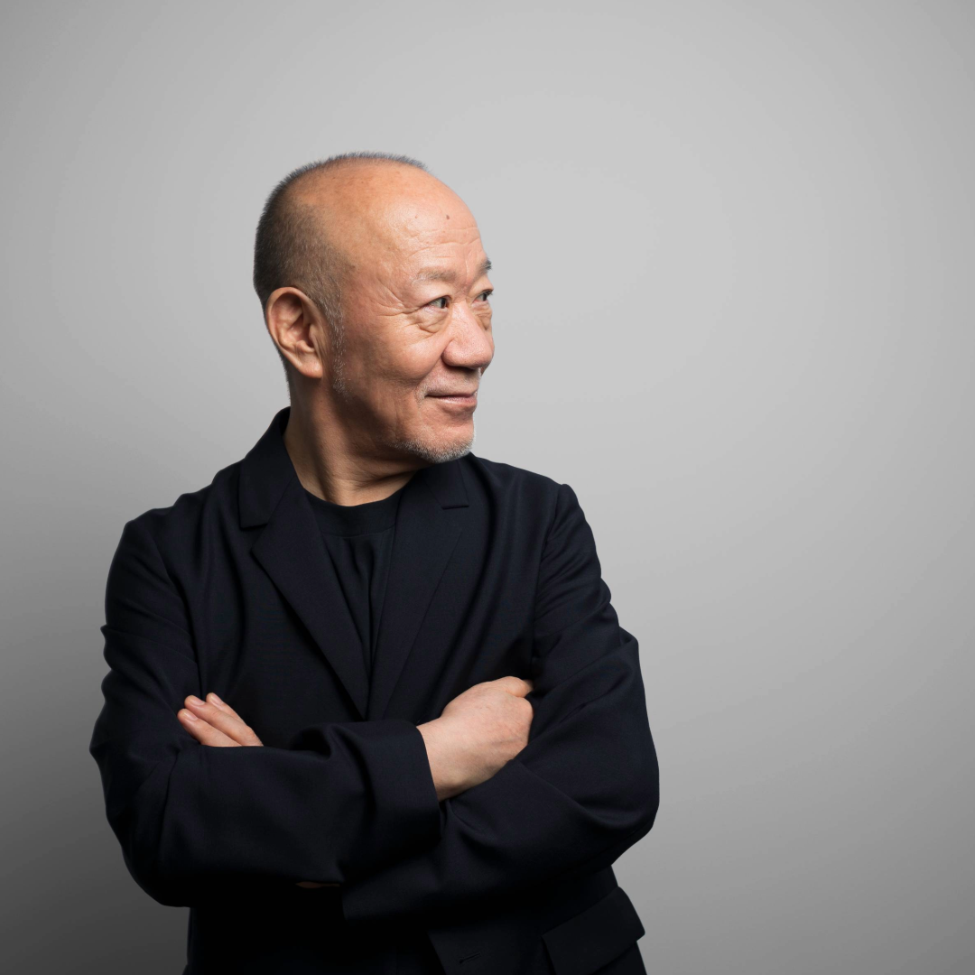 Older man in a dark blazer with arms crossed, looking to the side against a plain gray background