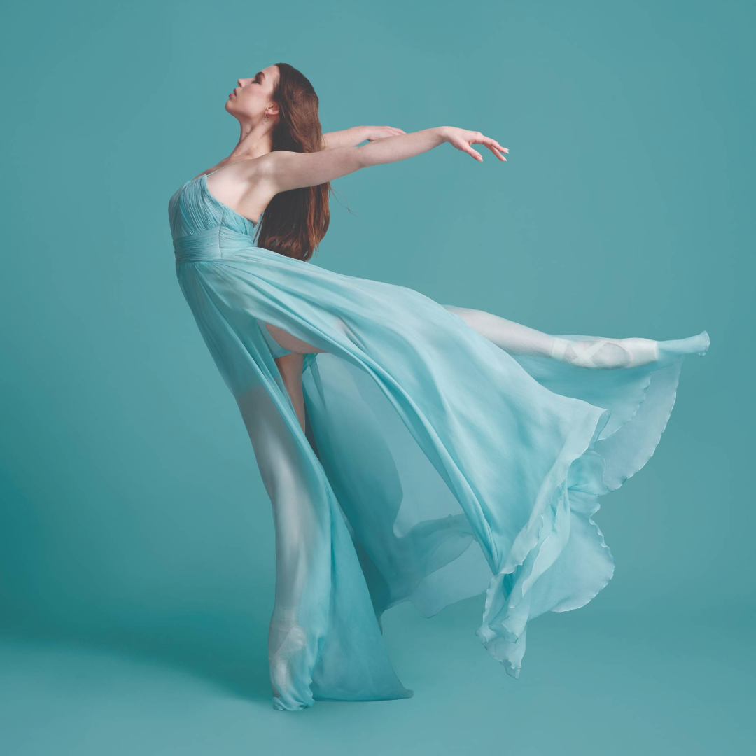 Dancer in a flowing light-blue dress performs an arched pose against a teal background, arms extended.