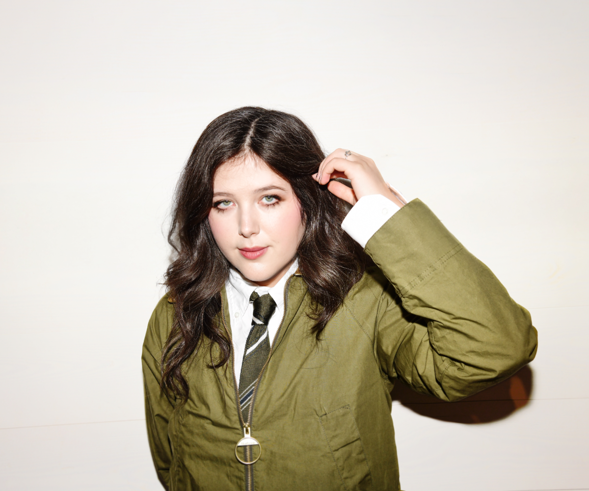 Woman with dark wavy hair posing with hand in hair, wearing an olive jacket and striped tie.