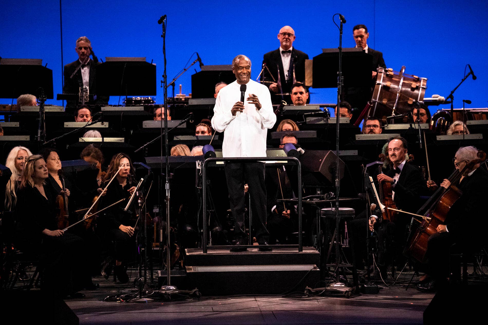 Thomas Wilkins white shirt stands at microphone, surrounded by orchestra with instruments and black music stands on stage