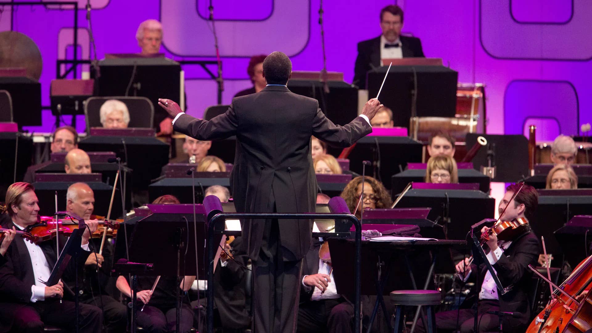 Conductor in a black suit stands with arms out before an orchestra onstage under purple lighting.