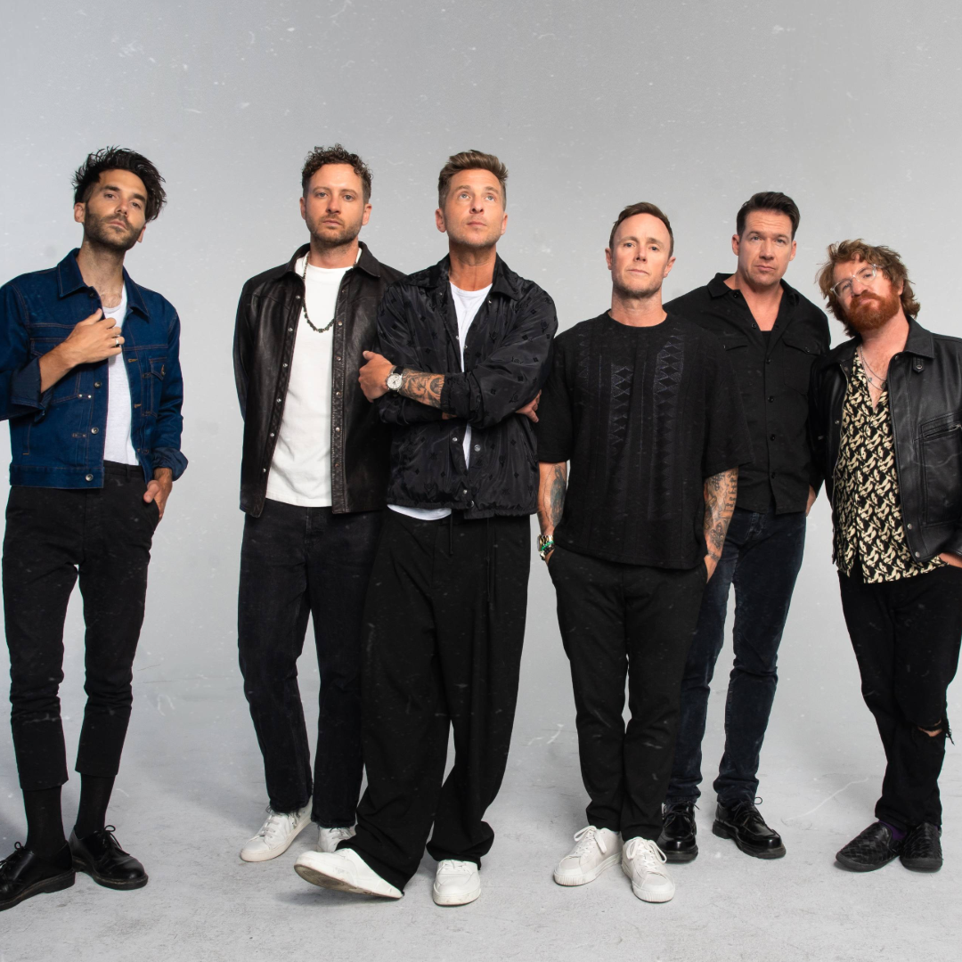 Six men posing together in casual dark outfits against a light gray studio backdrop.