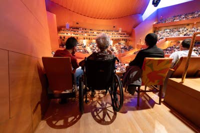 Audience seated in theater balconies, with people in wheelchairs watching a performance under colorful stage lighting.