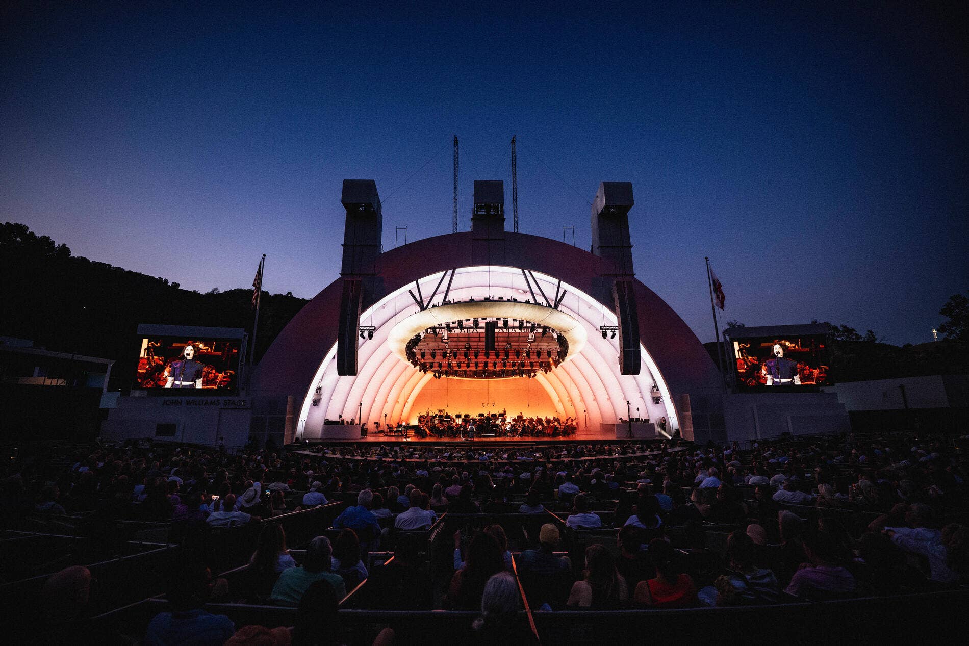 Hollywood Bowl at dusk with a lit stage, orchestra setup, and a crowd in the foreground
