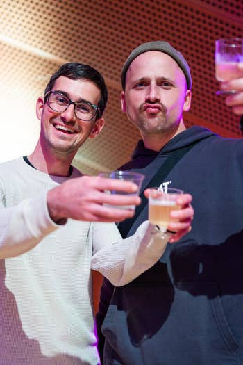 Two friends smiling and raising drinks at a party with purple and red lighting.