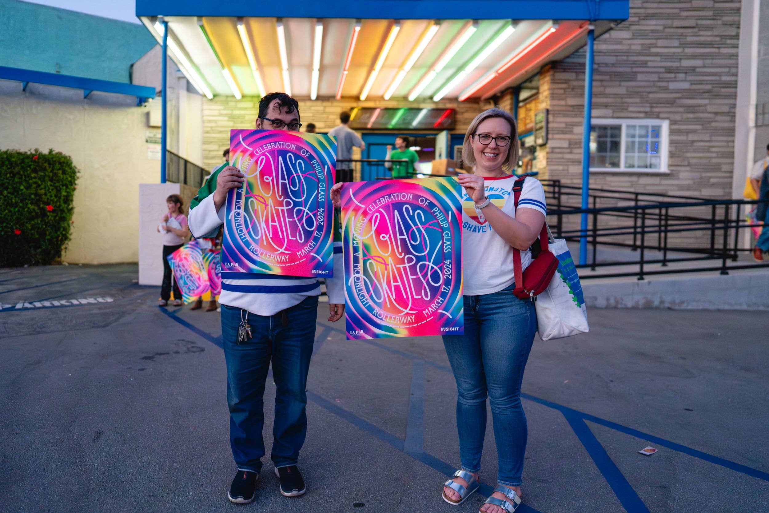 Two people hold colorful Glass Skates posters at an outdoor event under a brightly lit canopy.
