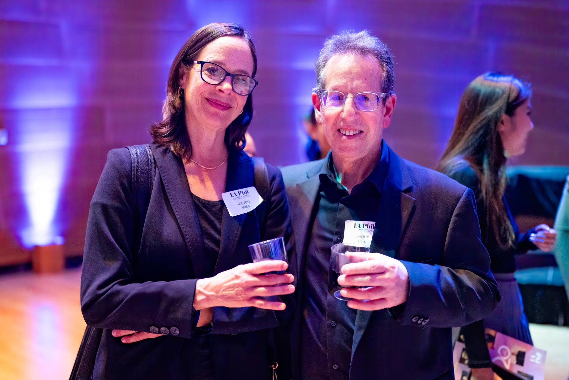 Two adults in suits and glasses smile at an event, holding wine glasses with purple-blue stage lighting behind them