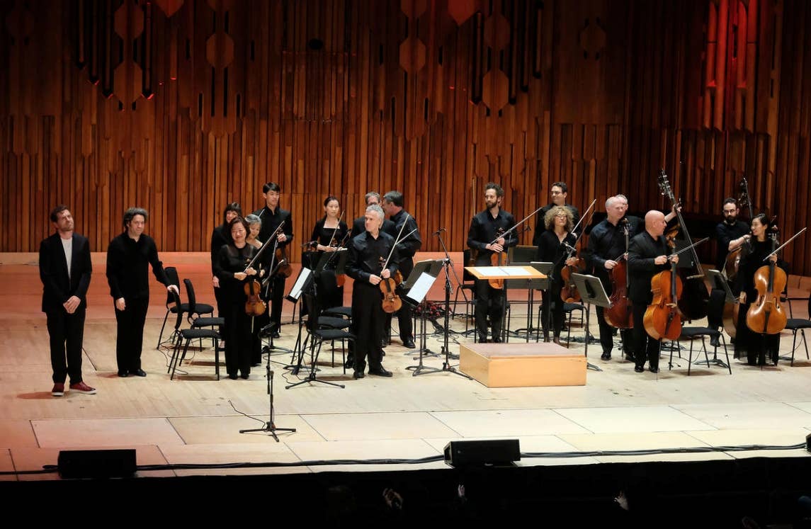 Orchestra ensemble in formal black attire performing on stage with violins, cellos, and wooden organ-like backdrop.