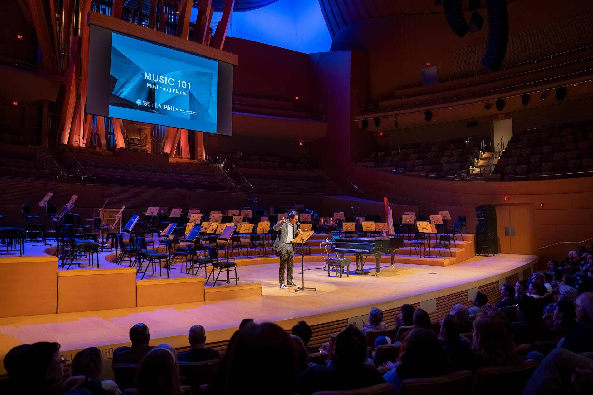 Musician stands at microphone on a concert hall stage with blue lighting and rows of empty music chairs.
