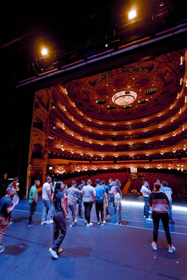 People in light-blue shirts walk across stage in an ornate theater with warm chandeliers and curved ceiling.