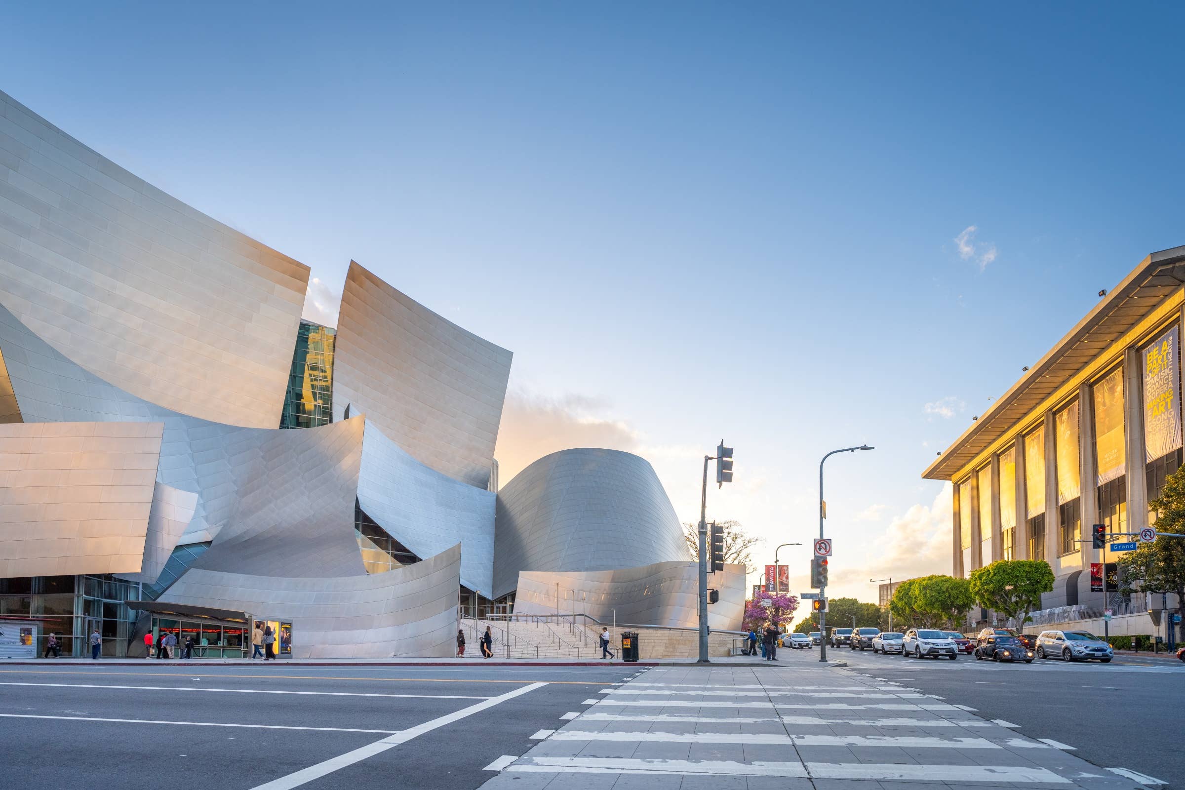 Modern museum buildings with curved white stone facade under a blue sky, street with cars and pedestrians.