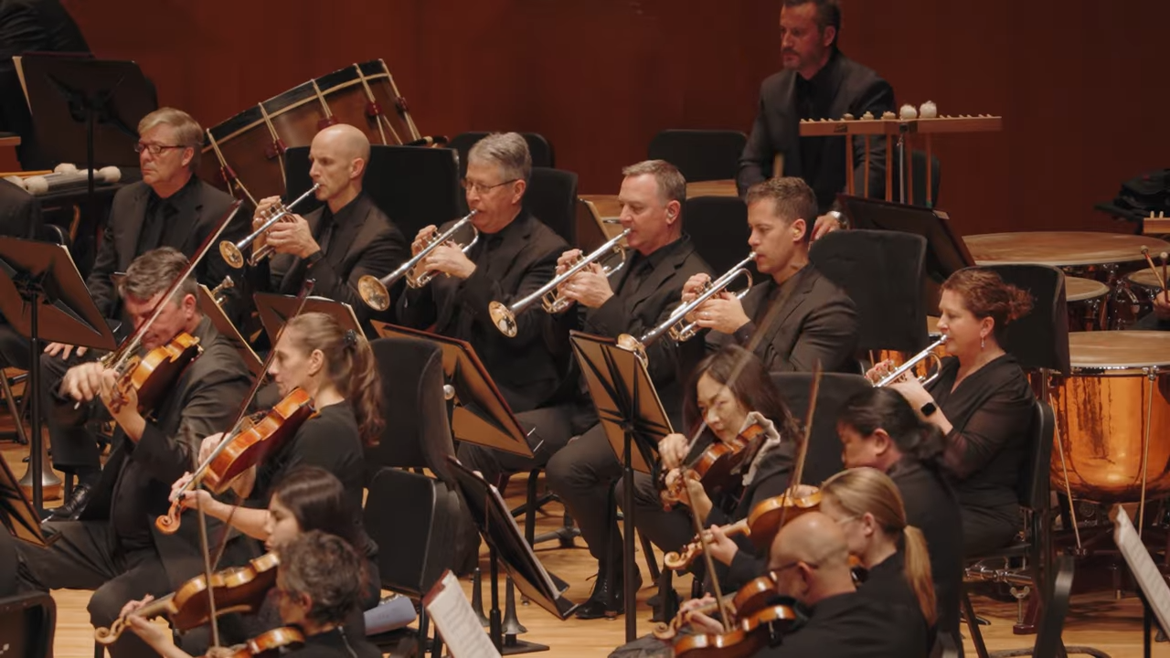Orchestra musicians in black performing with trumpets and strings on stage with music stands and instruments visible
