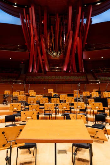 Empty concert hall with rows of wooden chairs, microphones, and red stage lighting behind a central pipe-like structure