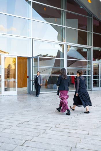 People walk outside modern glass building with curved roof and interior lights visible through windows.