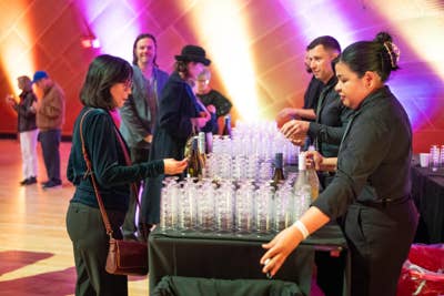 Two women pour and chat at a party table of sparkling drinks amid colorful stage lighting and guests in the background