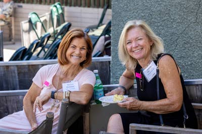Two smiling women seated outdoors at an event table with badges, drinks, and chairs behind them