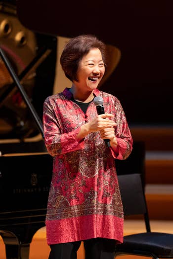 Smiling woman sings into a handheld microphone on stage, with a grand piano and dark backdrop behind her.